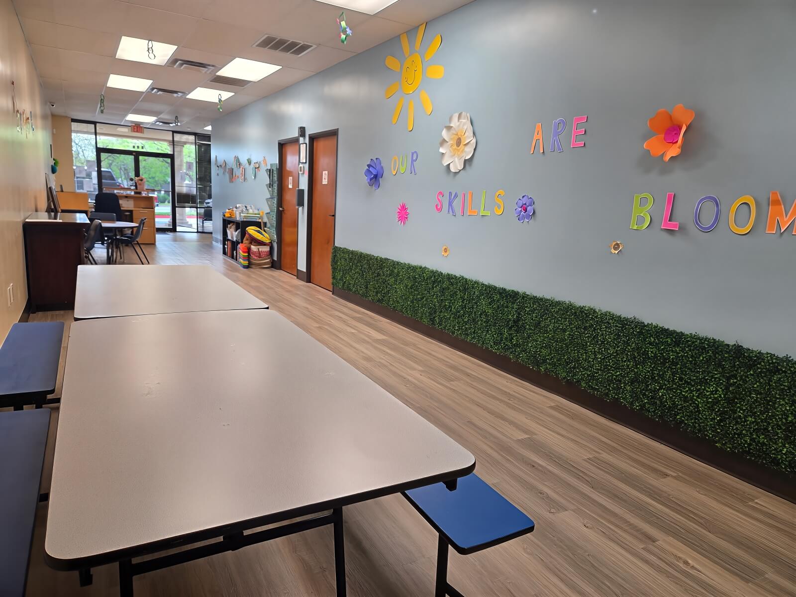 CAI ABA therapy classroom in Hurst TX with learning tables benches and Our Skills Are Blooming wall display featuring handmade flowers and sunshine decorations